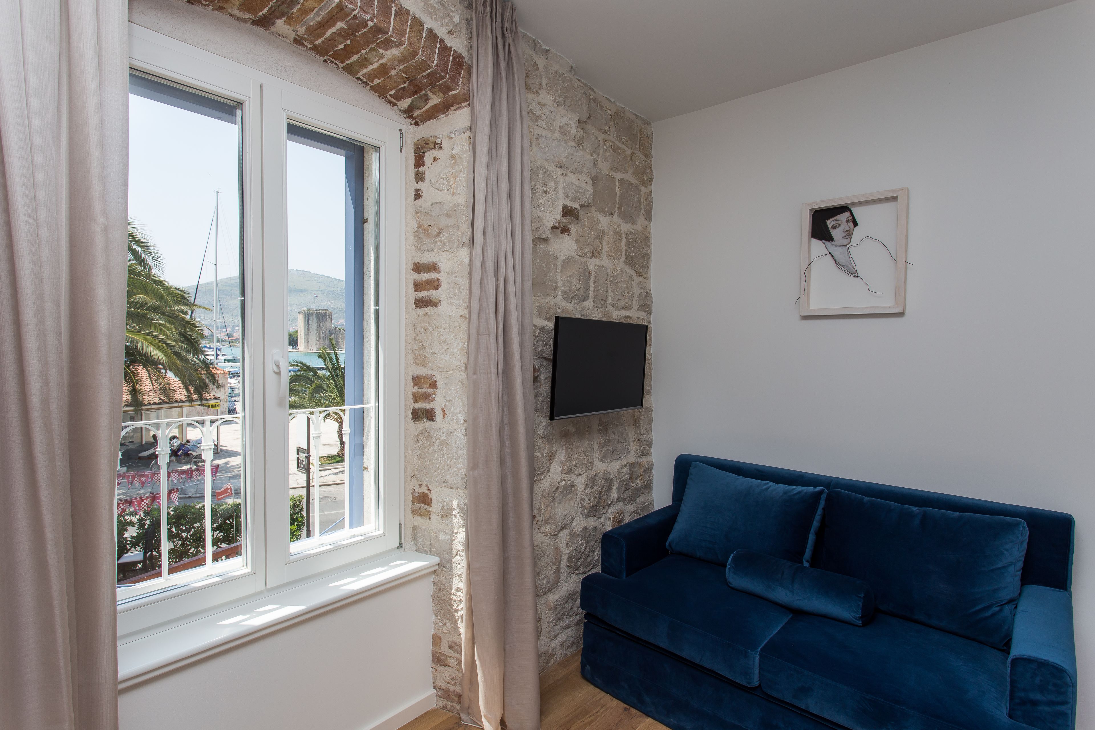 A room with a stone wall, a window with curtains, a blue sofa, and a flat-screen TV mounted on the wall.