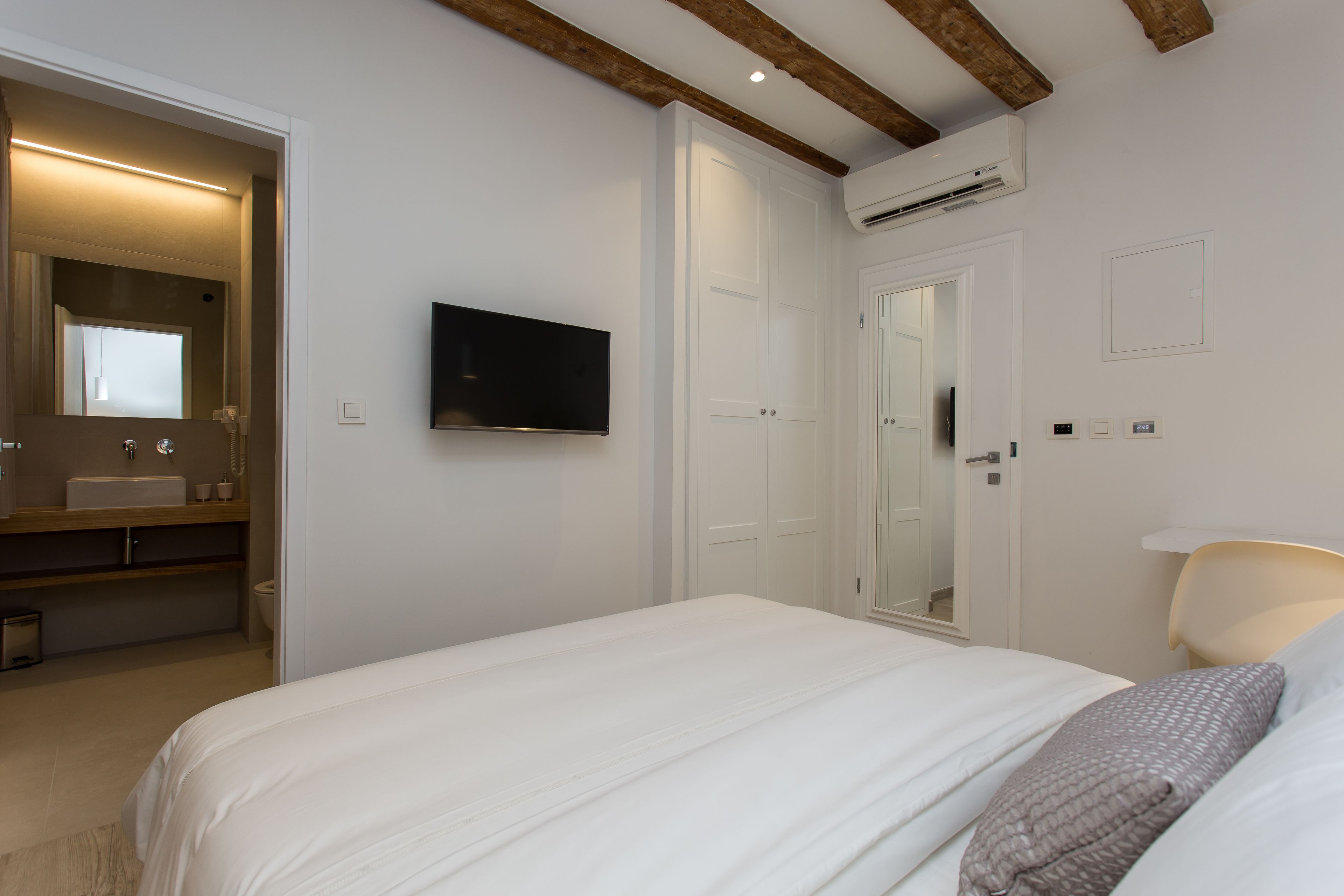 A bedroom with a bed, a TV, a mirror, and a bathroom with a sink and toilet.
