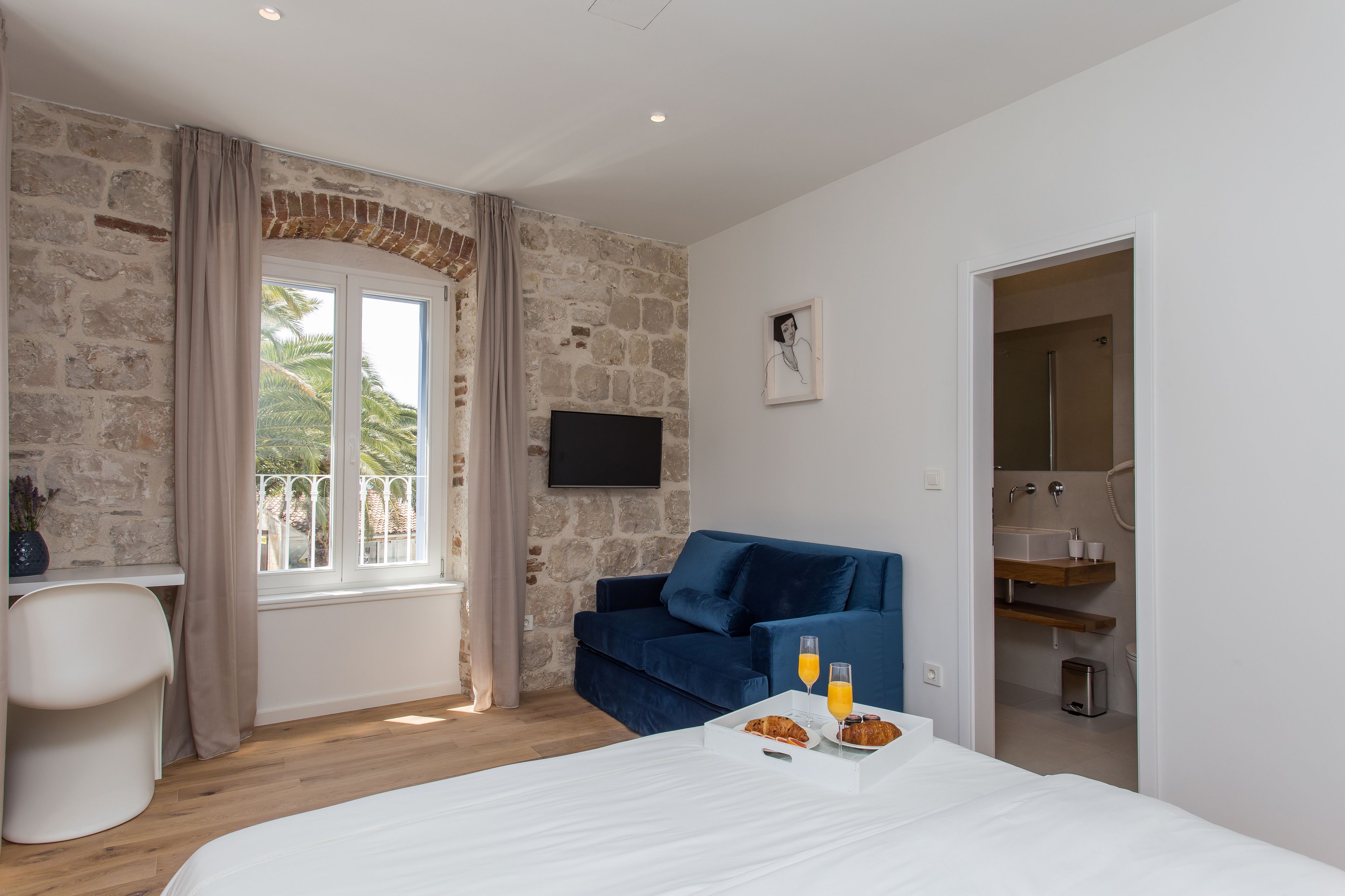 Habitación de hotel moderna con pared de piedra, sofá azul, televisor de pantalla plana, bandeja de desayuno y vistas a palmeras.