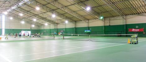 Pista de tenis interior con varias canchas, una red y una pared verde con logotipos de patrocinadores.
