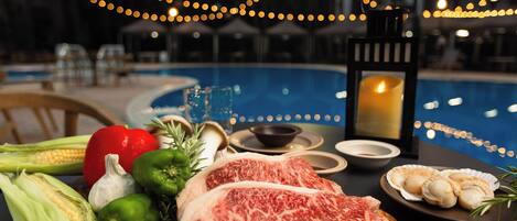 A dining table with steak, vegetables, and seafood, a candle, and a poolside setting.