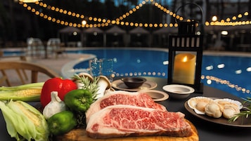 A dining table with steak, vegetables, and seafood, a candle, and a poolside setting.