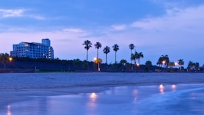A beach with a hotel in the background, palm trees, and lights reflecting on the water.