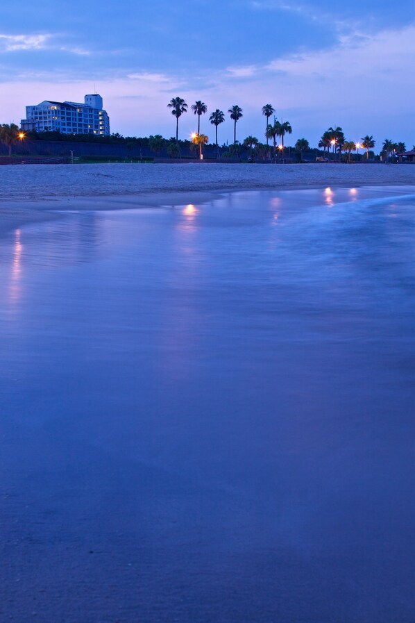 Une plage avec un hôtel en arrière-plan, des palmiers et des lumières qui se reflètent sur l’eau.