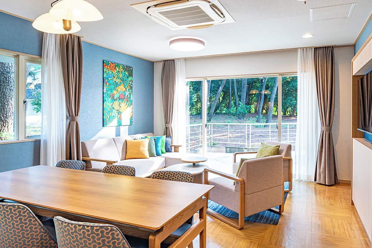 A modern hotel room with a wooden dining table, patterned chairs, a sofa, and a large window offering a view of the outdoors.
