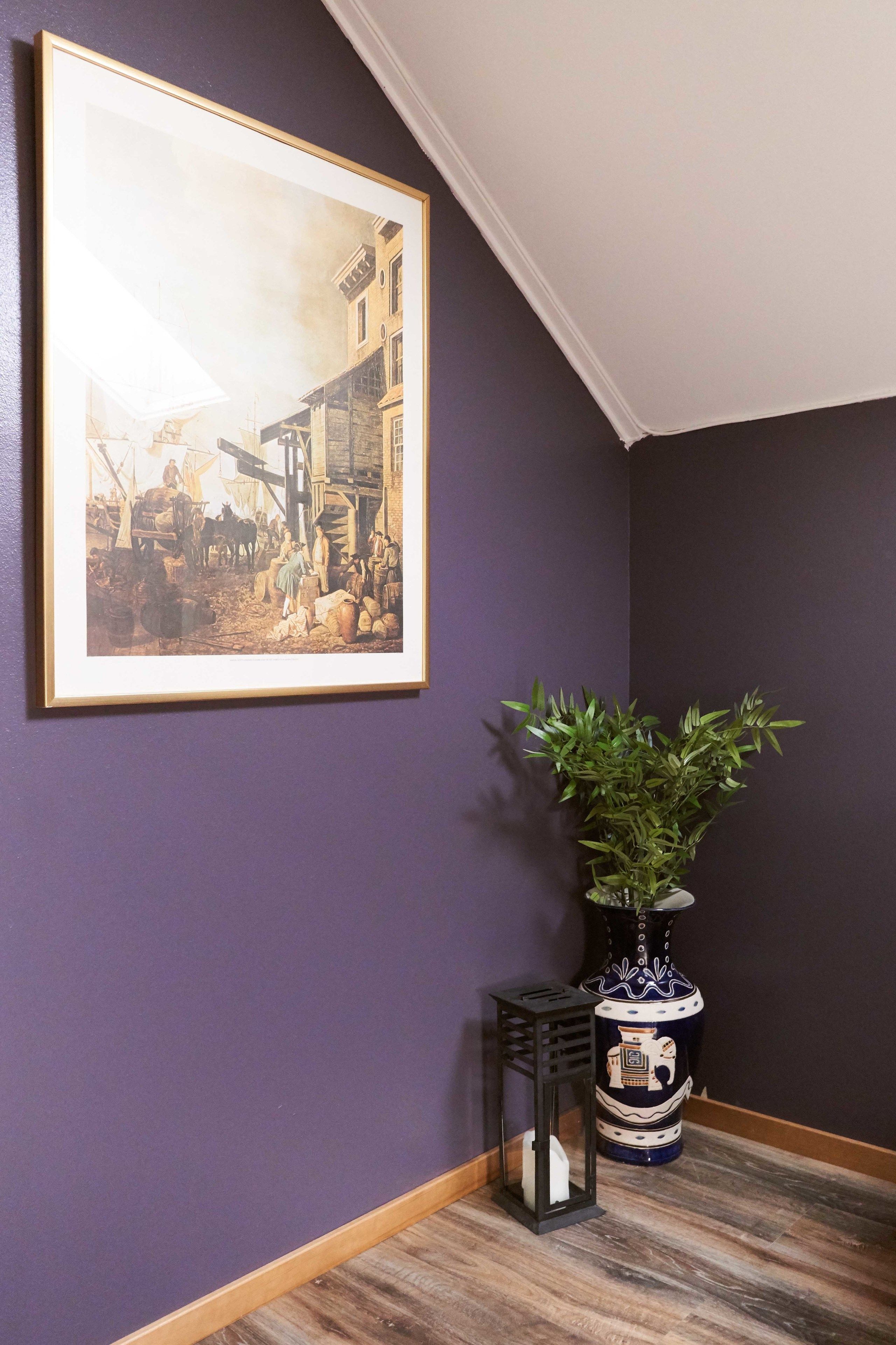 Una habitación con pared morada, un cuadro histórico enmarco, una planta en maceta y un jarrón decorativo.
