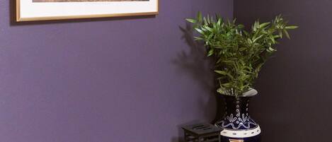 Una habitación con pared morada, un cuadro histórico enmarco, una planta en maceta y un jarrón decorativo.