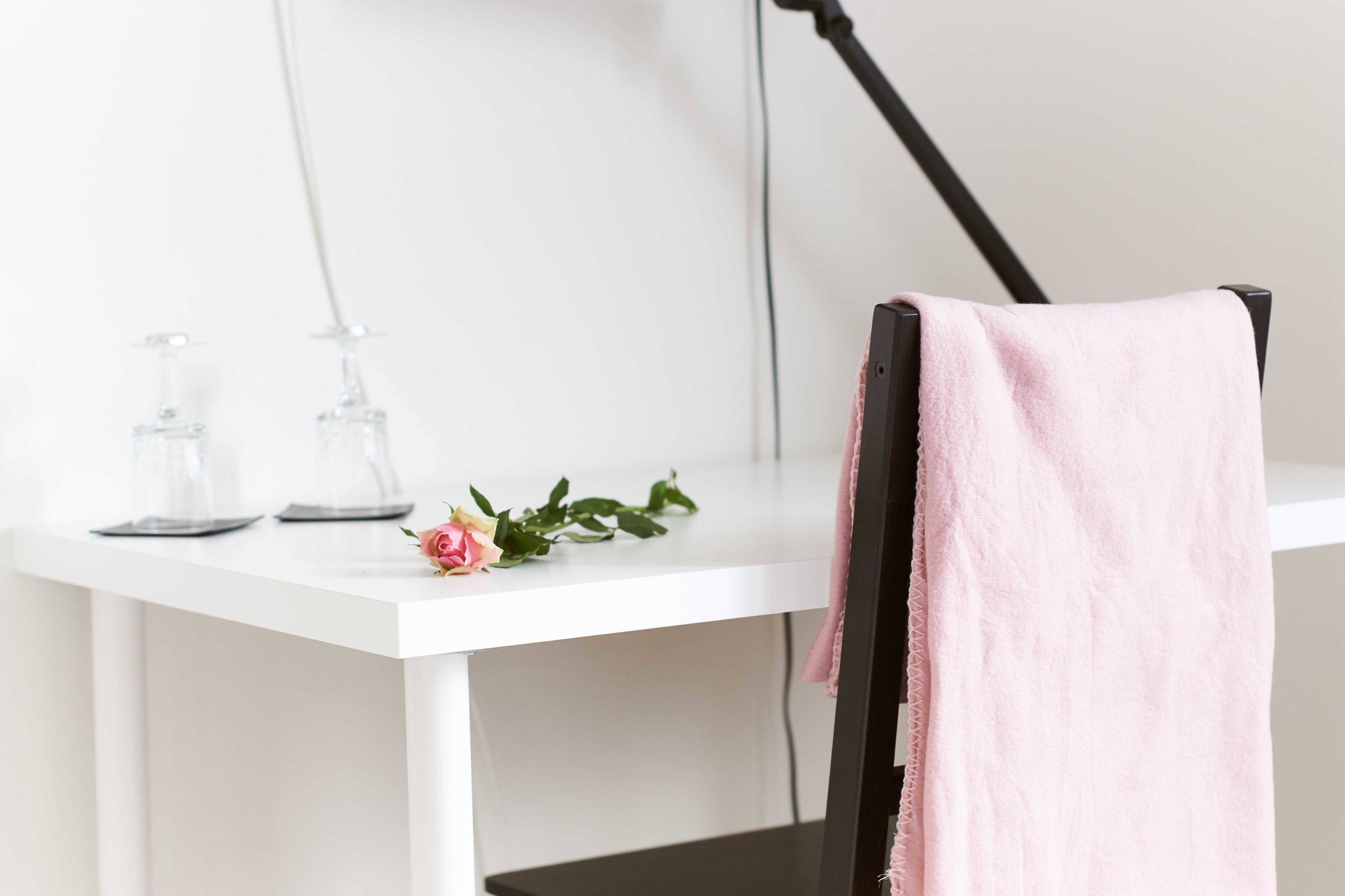Una scrivania bianca con un asciugamano rosa appoggiato su una sedia, una bottiglia di vetro e una rosa sulla scrivania.