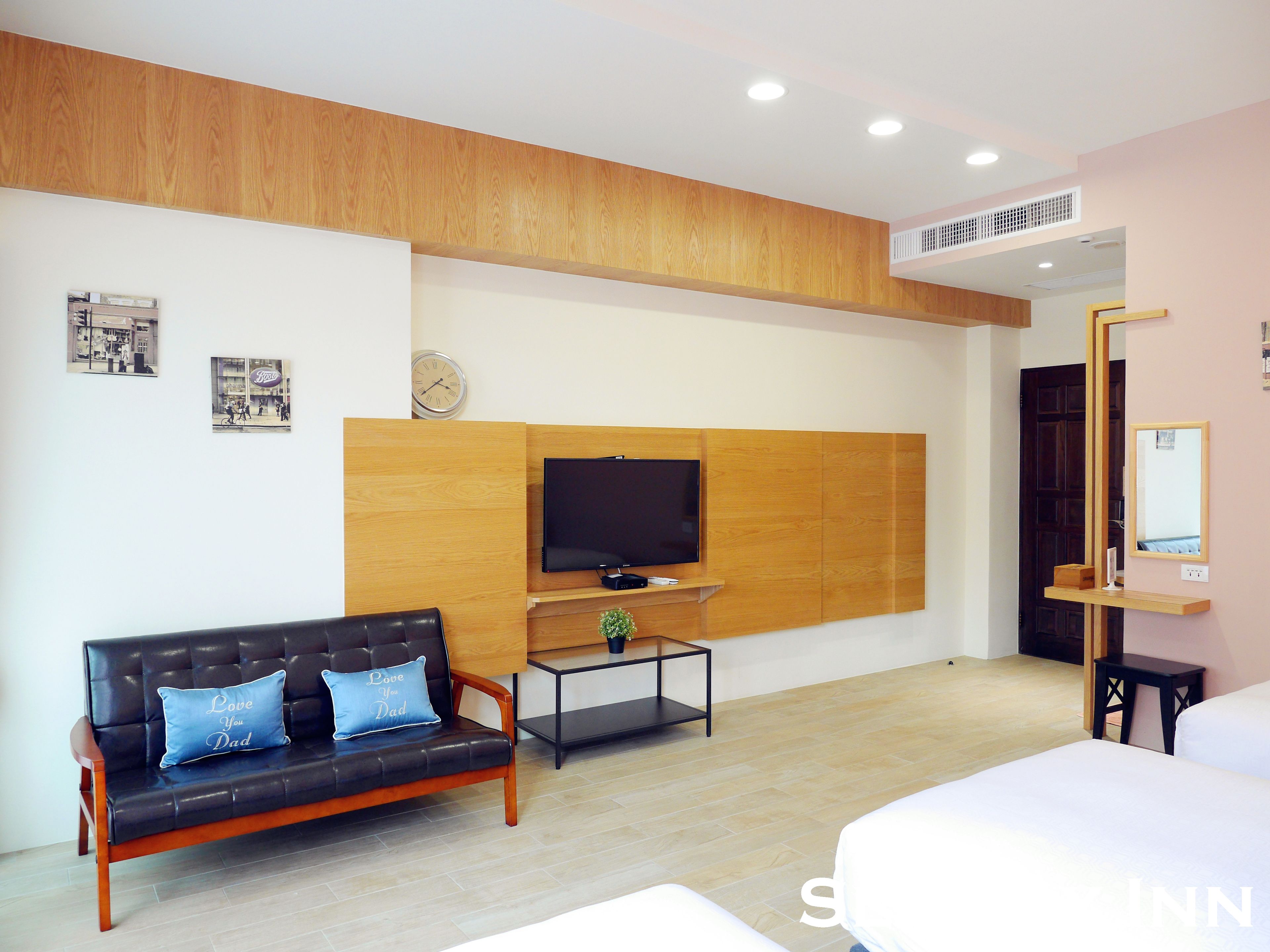 A modern hotel room with a wooden accent wall, a flat-screen TV, a sofa with decorative pillows, a bedside table, and a large bed.