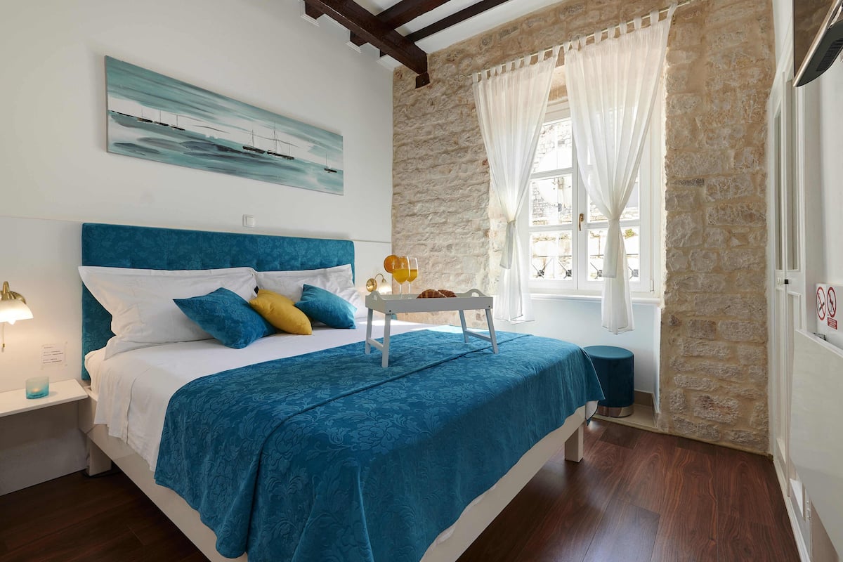 A bedroom with a stone wall, a bed with a blue headboard, a nightstand, a bedside table, a window with curtains, and a painting on the wall.