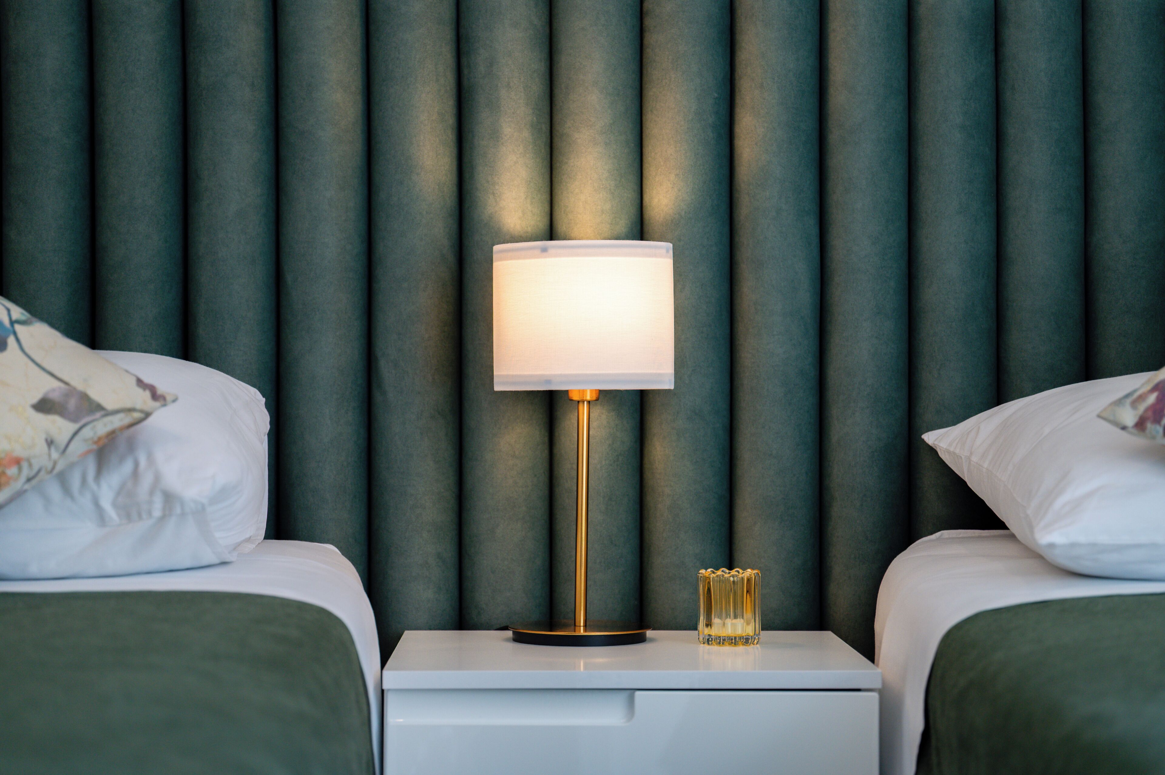 A hotel room with two beds, a bedside table with a lamp, and a green upholstered headboard.
