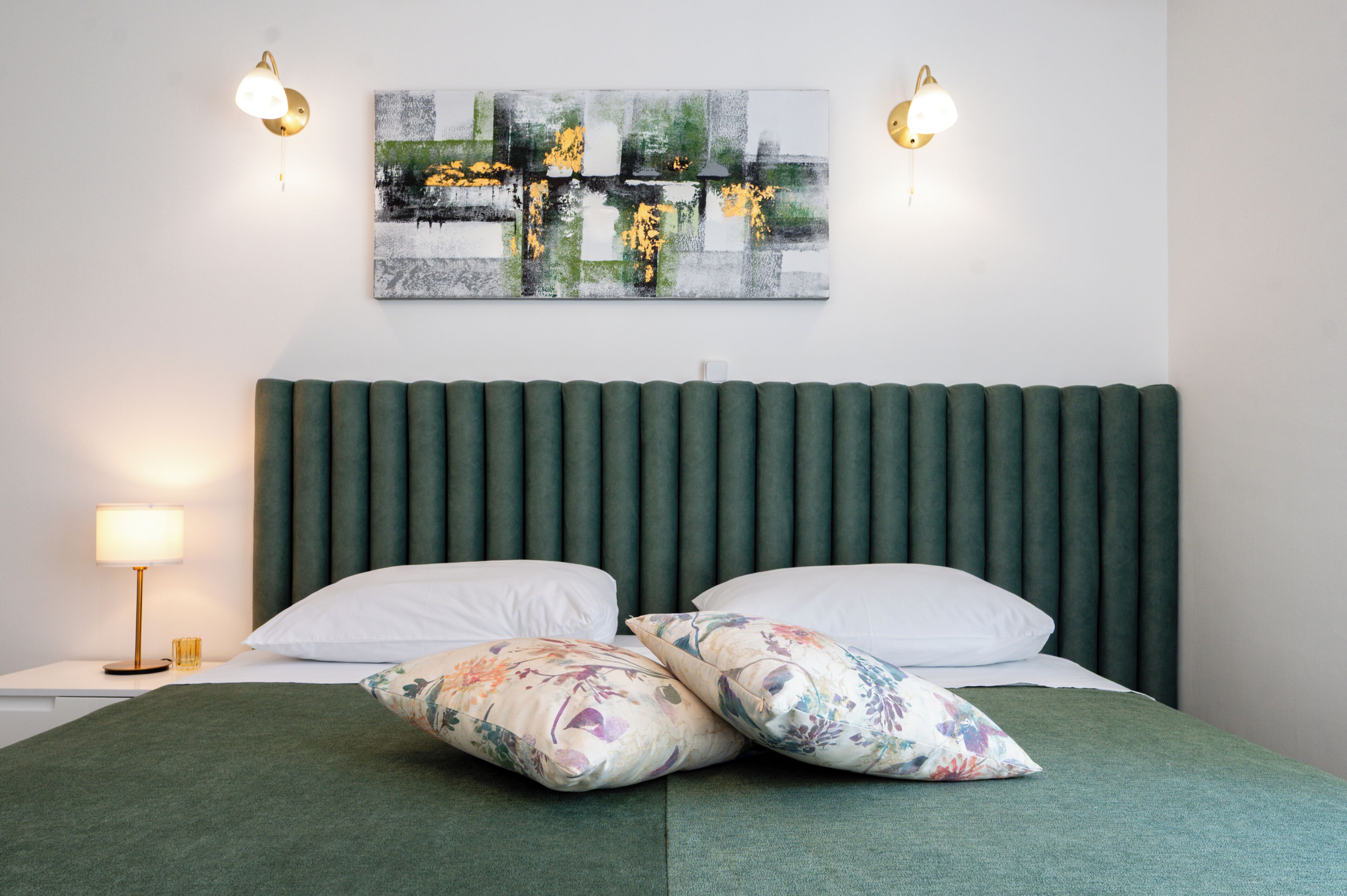 A bed with green bedding, two white pillows, and a green headboard. There is a wall-mounted abstract painting and two wall sconces.
