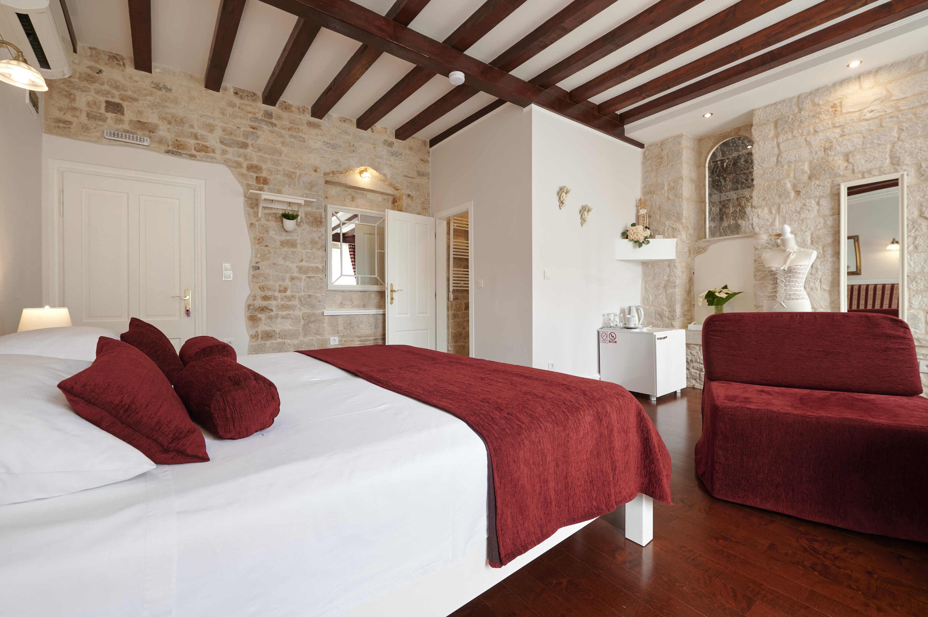A bedroom with a stone wall, wooden beams, a bed with white and red bedding, a white bedside table, and a red chair.