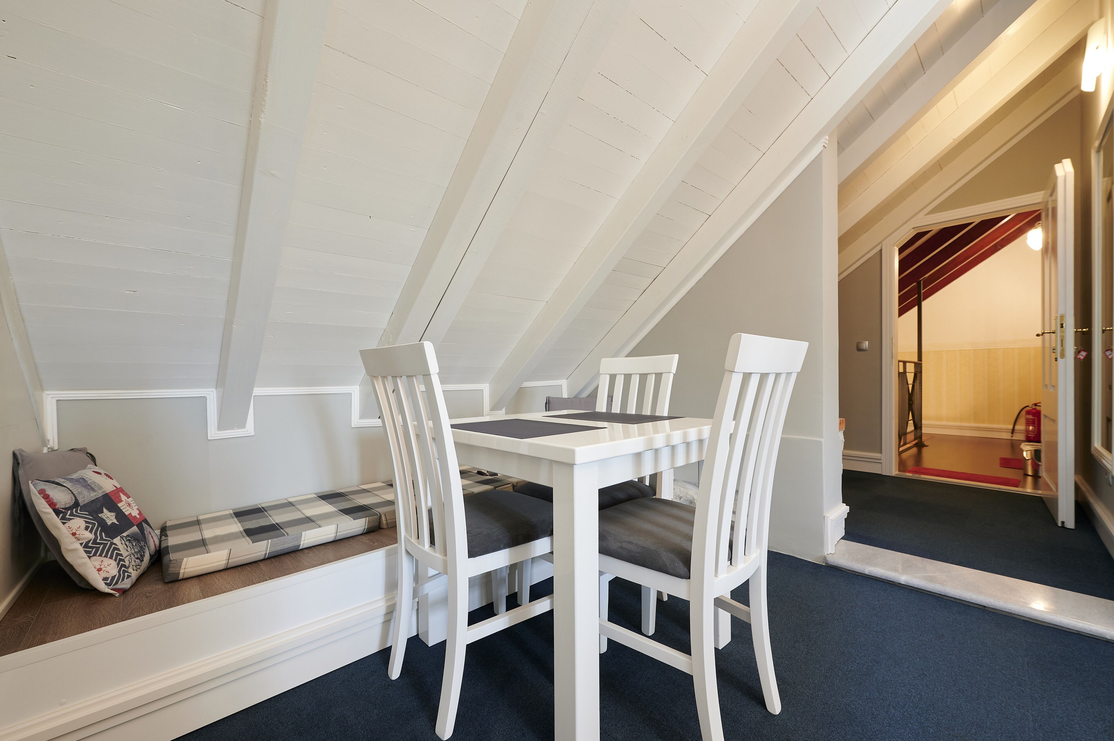 A dining area with a white table and chairs, a bench with cushions, and a view of a hallway with a red carpet.