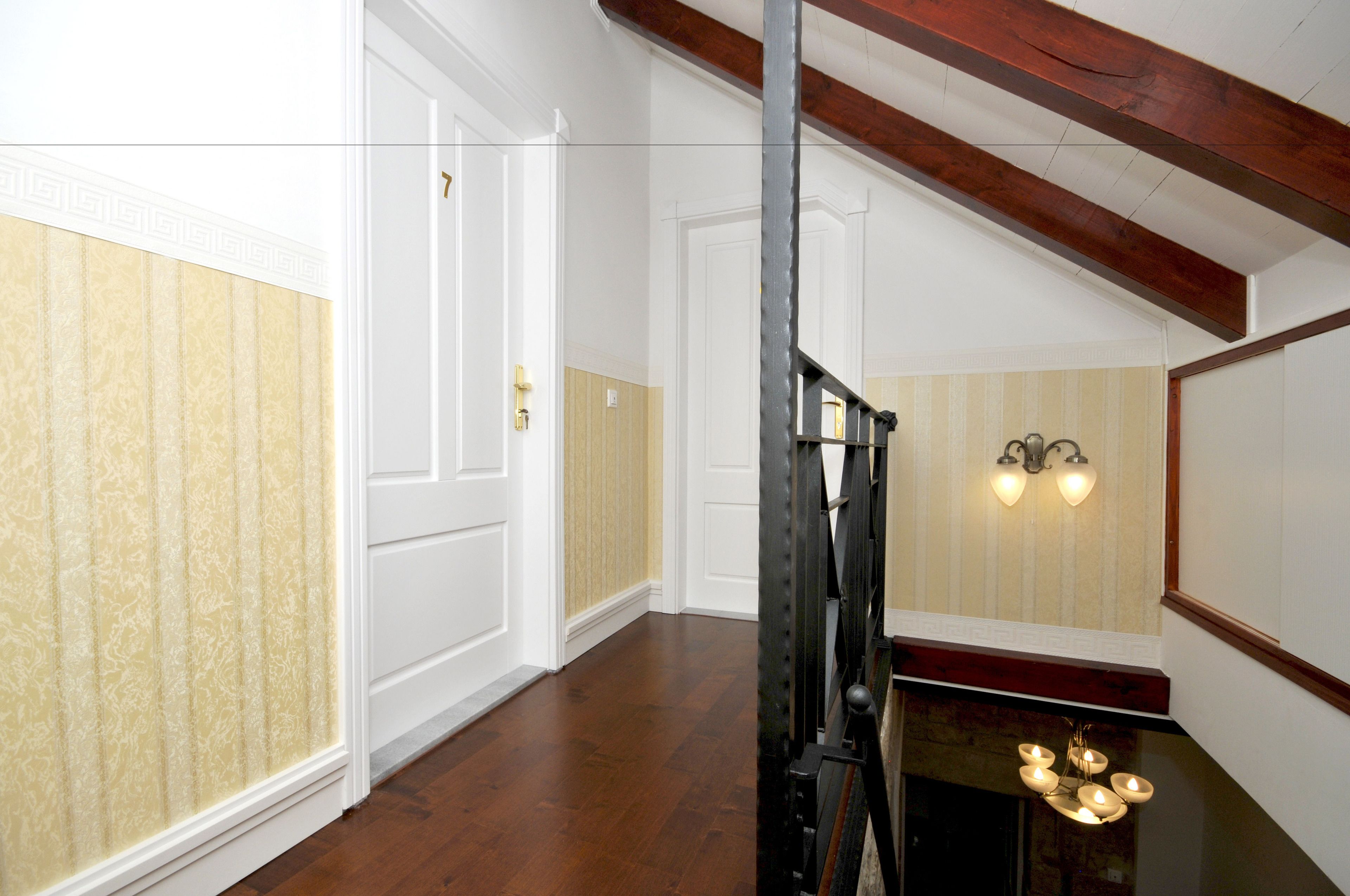 A hallway with wooden floors, white walls, and a staircase with a wooden railing. There are two wall sconces with lights on.