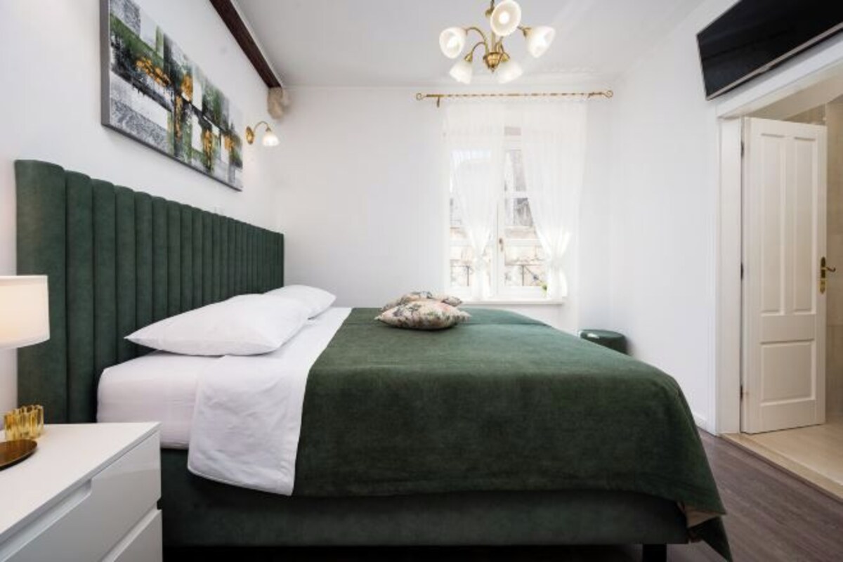 A bedroom with a green upholstered headboard, a white bedspread, a single pillow, a bedside table with a lamp, and a window with sheer curtains.