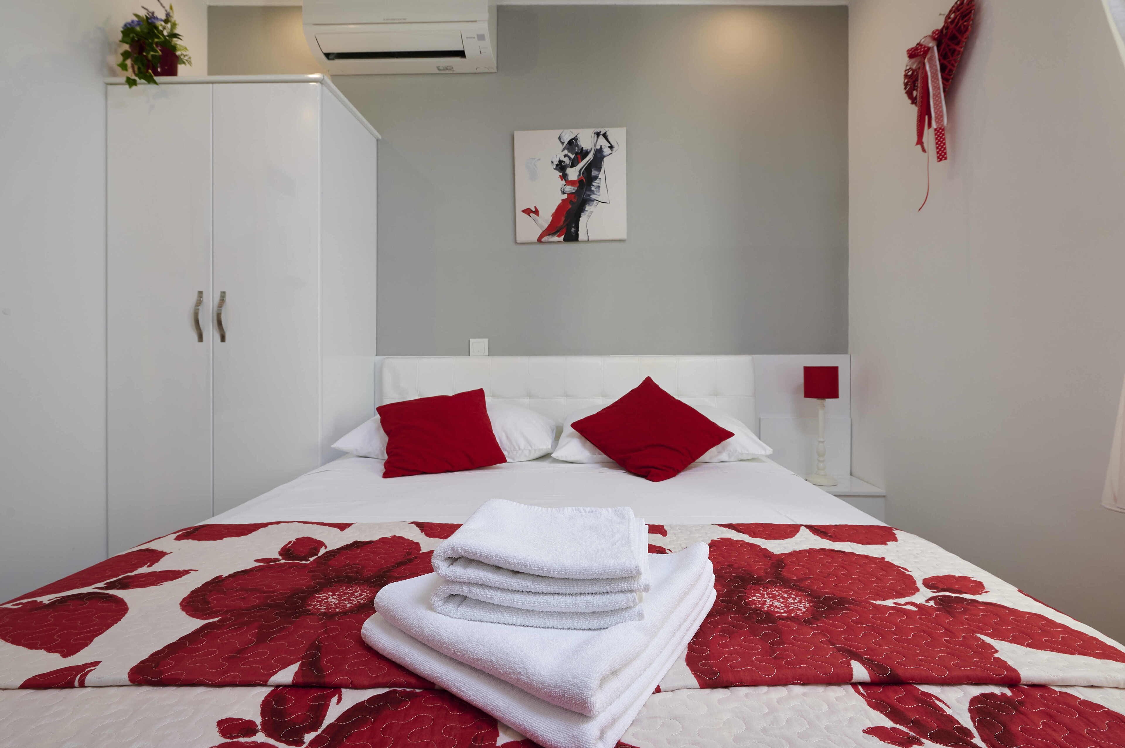 A neatly made bed with a red and white floral bedspread, a stack of white towels, and two red pillows.