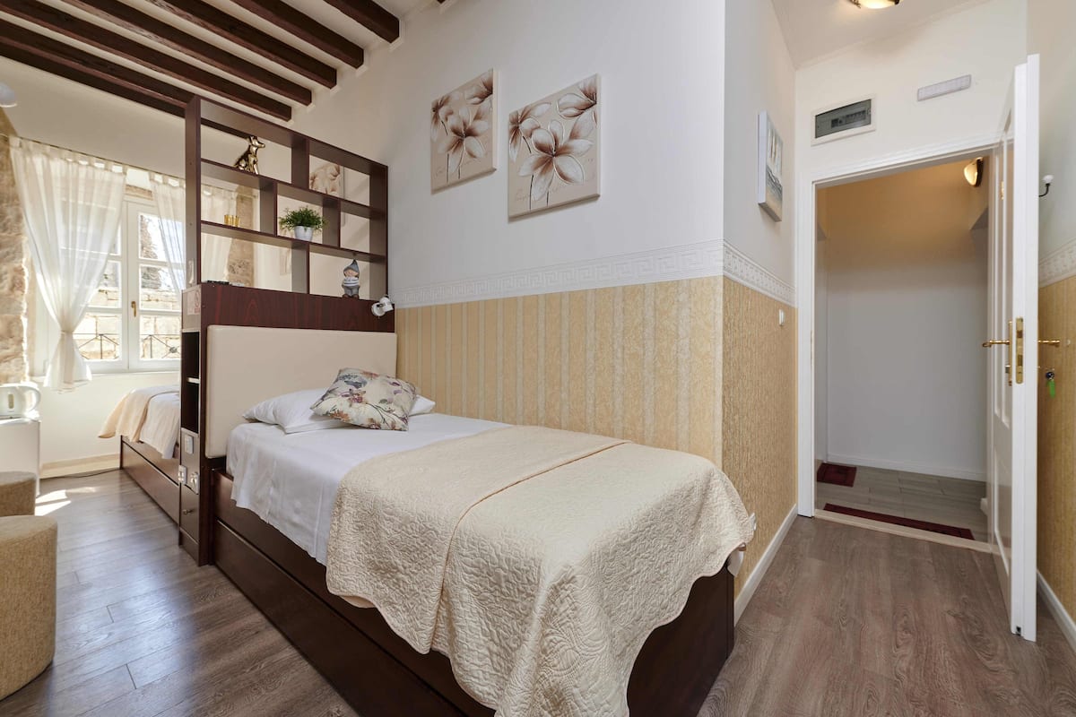A bedroom with a bed, a nightstand, a dresser, a window with curtains, and a door leading to another room.
