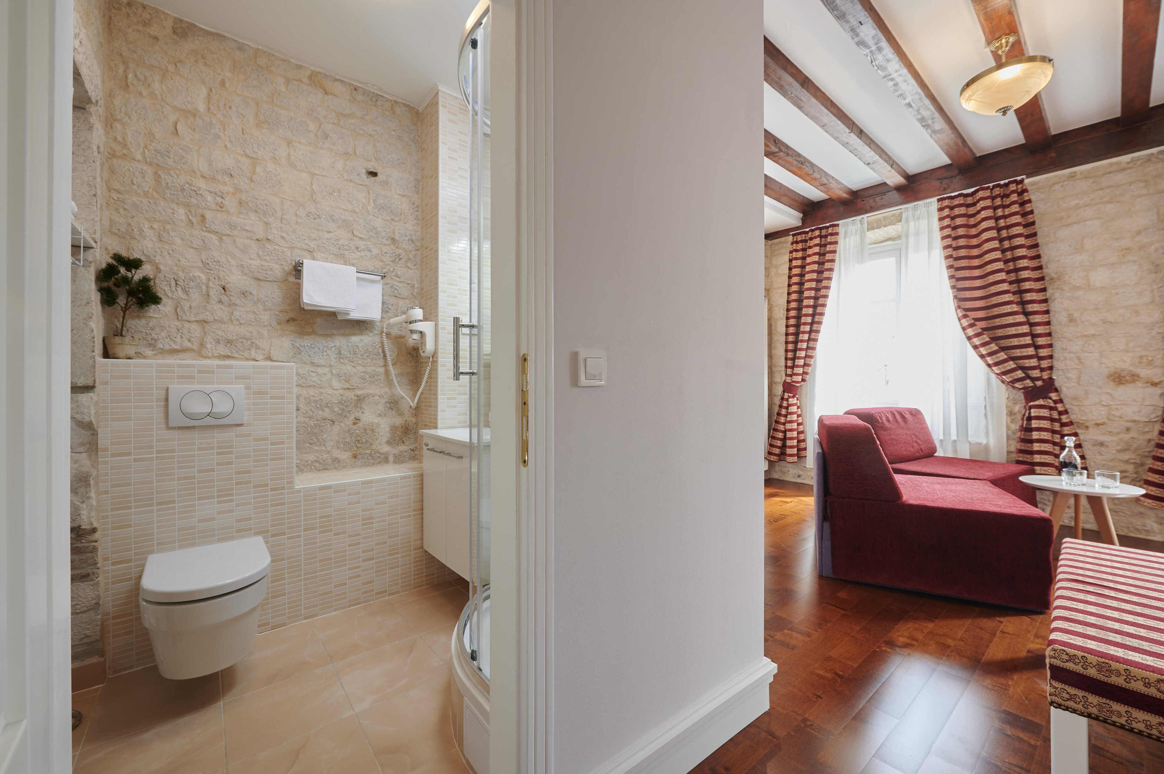 A bathroom with a toilet, shower, and a glass-enclosed shower stall. There is a red sofa and a small table with a glass and a bottle.
