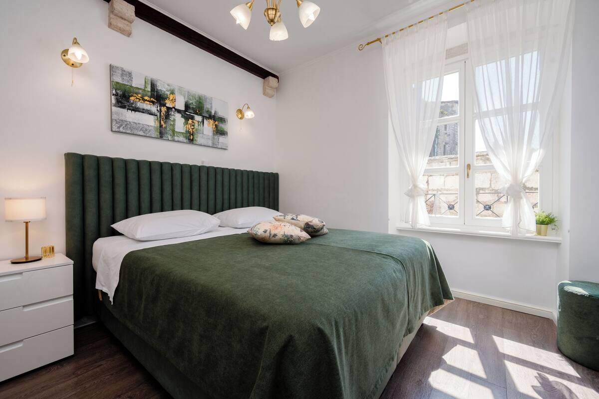 A neatly arranged bedroom with a bed, bedside table, lamp, artwork, and a window with curtains.