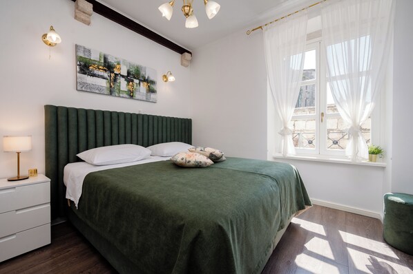 A neatly arranged bedroom with a bed, bedside table, lamp, artwork, and a window with curtains.