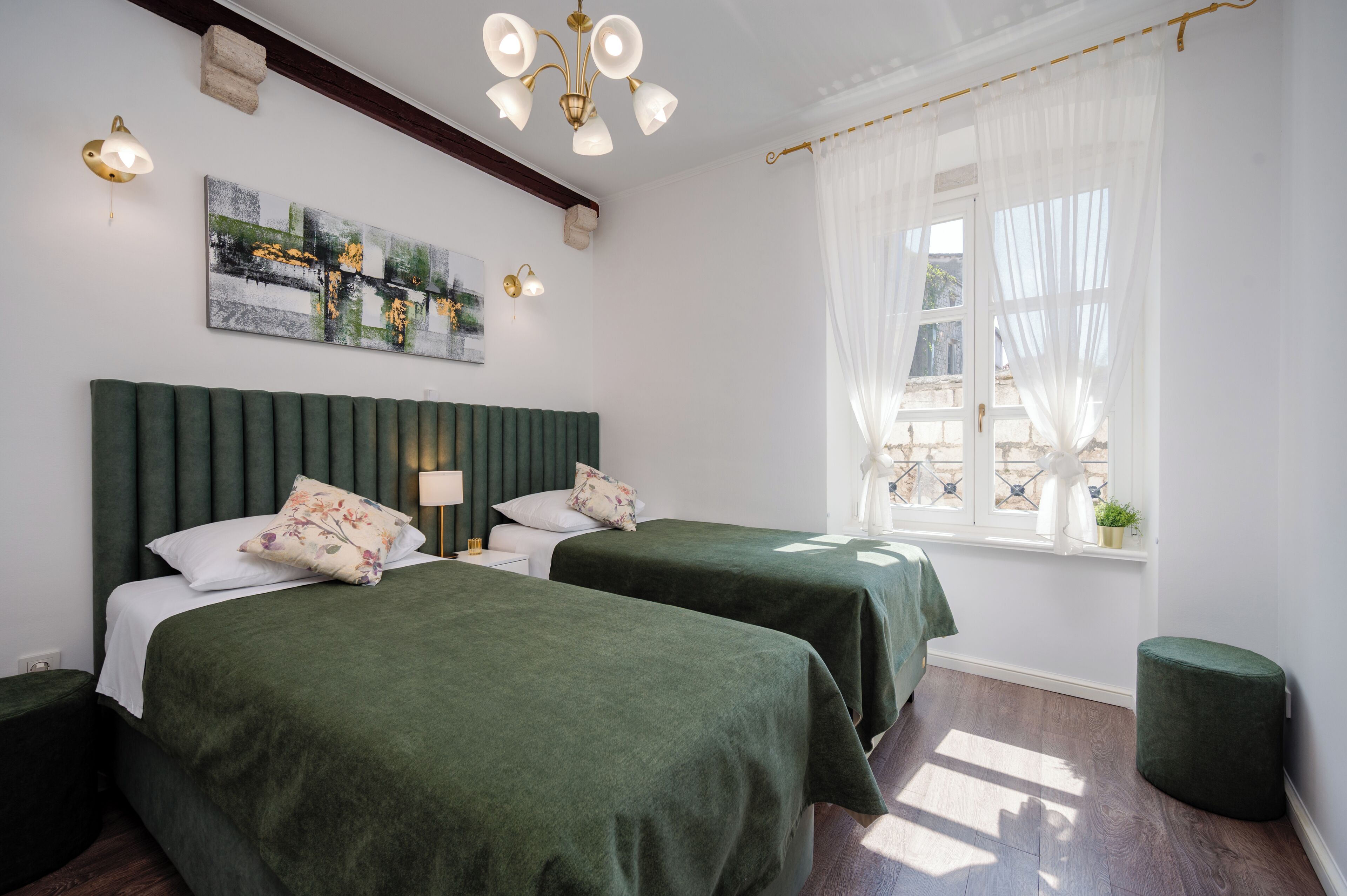 Habitación de hotel con dos camas, un ventanal grande con cortinas traslúcidas, un cabecero tapizado en verde y una lámpara fijada a la pared.