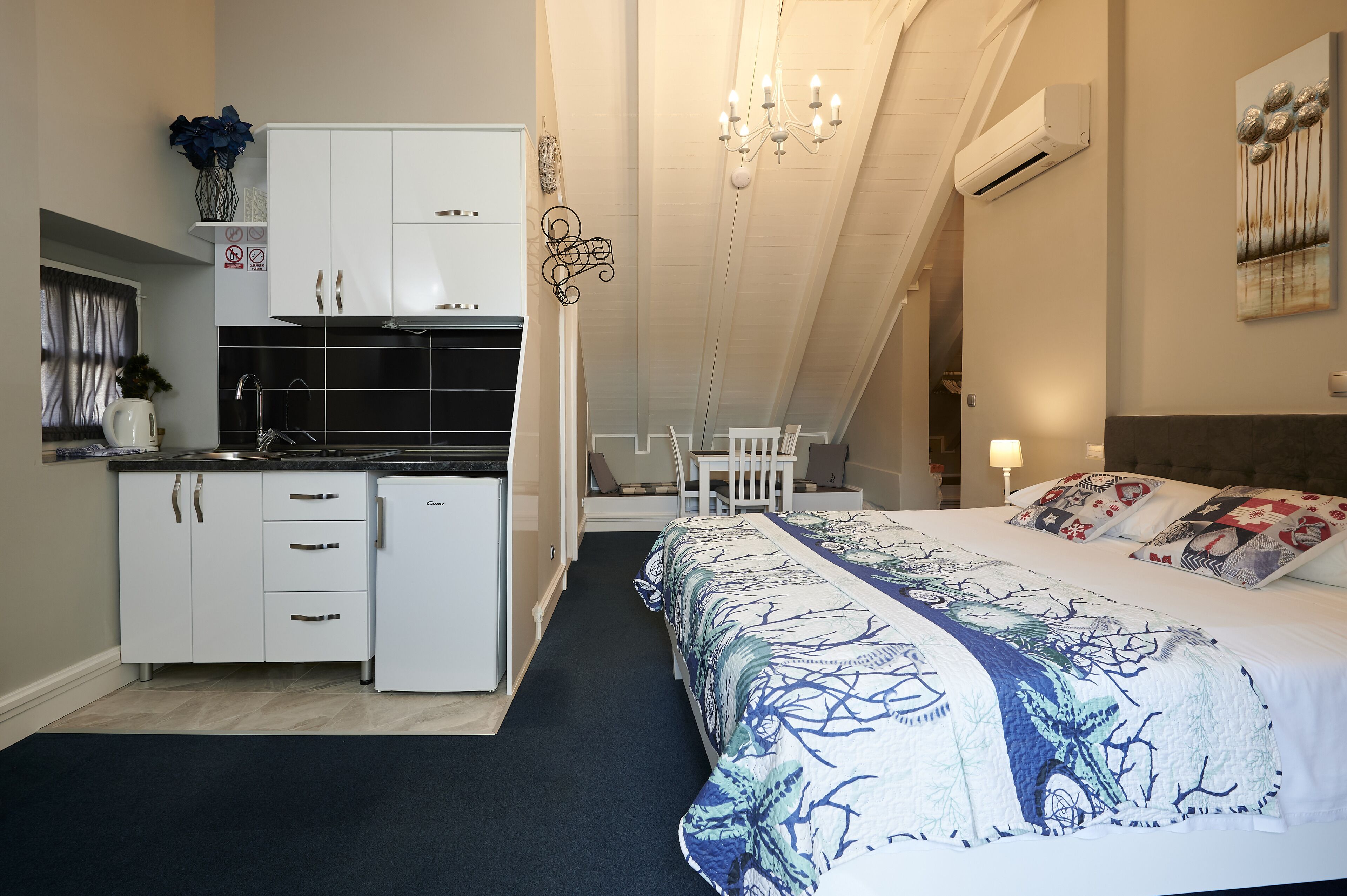 A compact hotel room with a kitchenette, a bed with patterned bedding, and a chandelier.