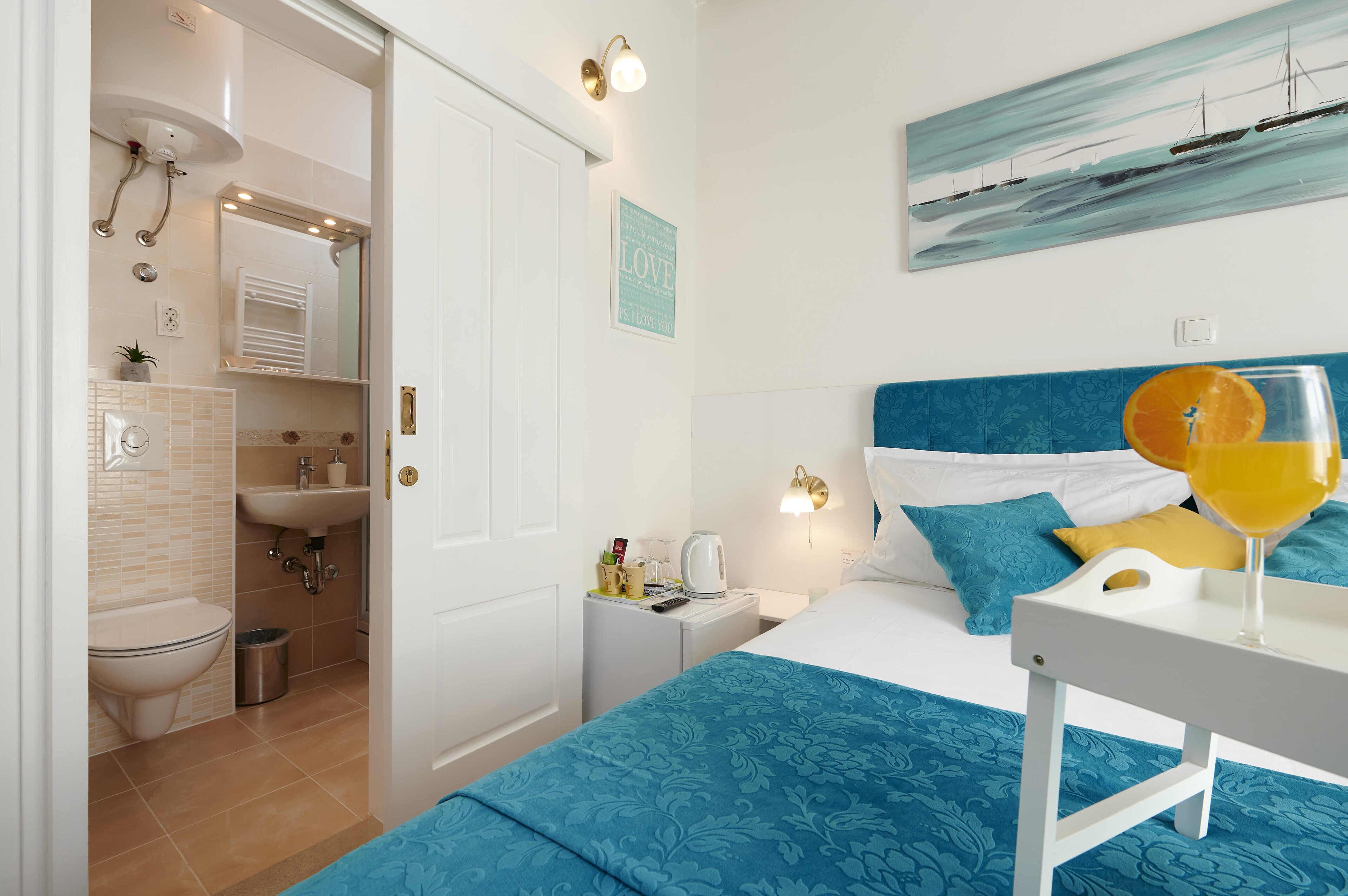 Una habitación de hotel con baño, una cama con cabecera azul, un cuadro con barcos y un vaso de zumo de naranja sobre la mesita de noche.