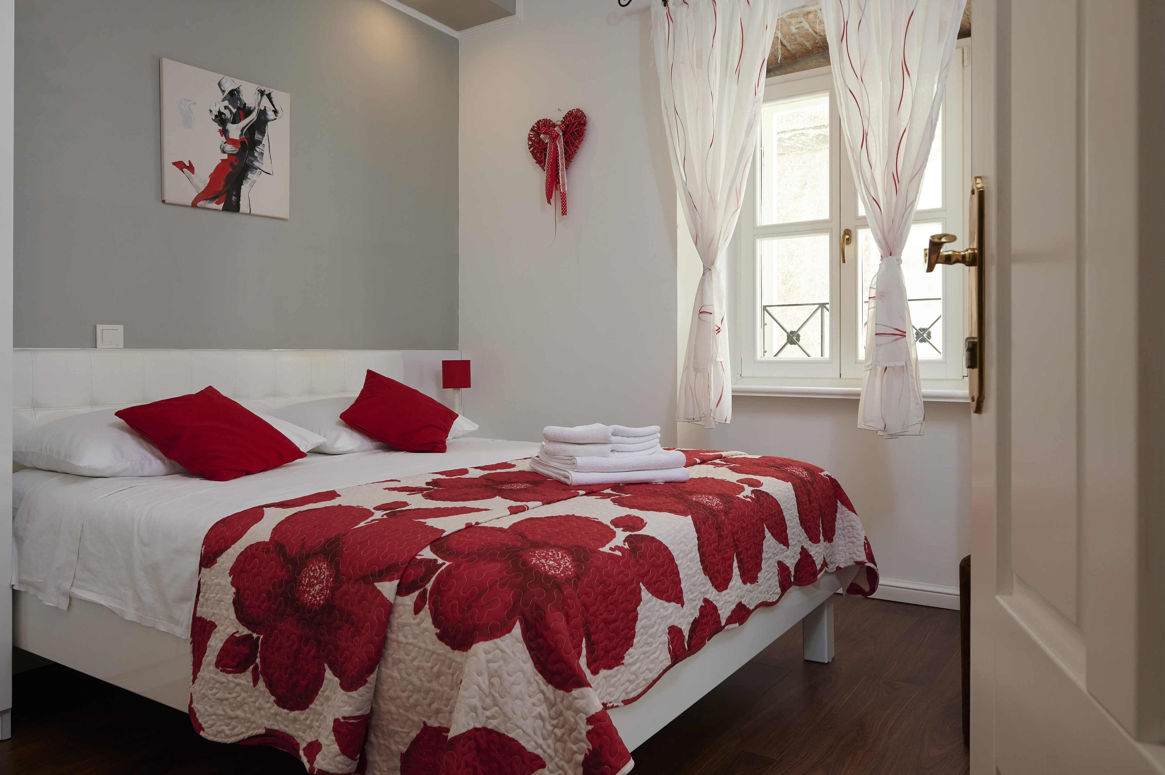 Una cama bien tendida con ropa de cama roja y blanca, una decoración con forma de corazón en la pared y una ventana con cortinas.