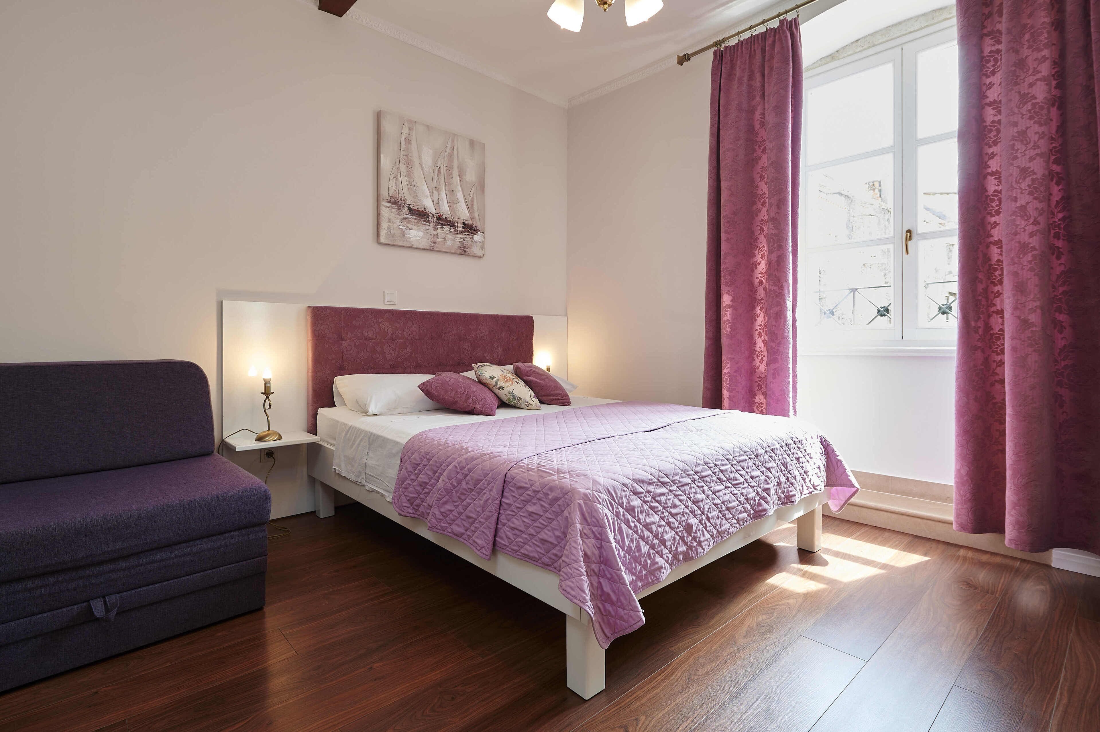 A bedroom with a bed, a nightstand, a bedside lamp, a window with curtains, and a sofa.