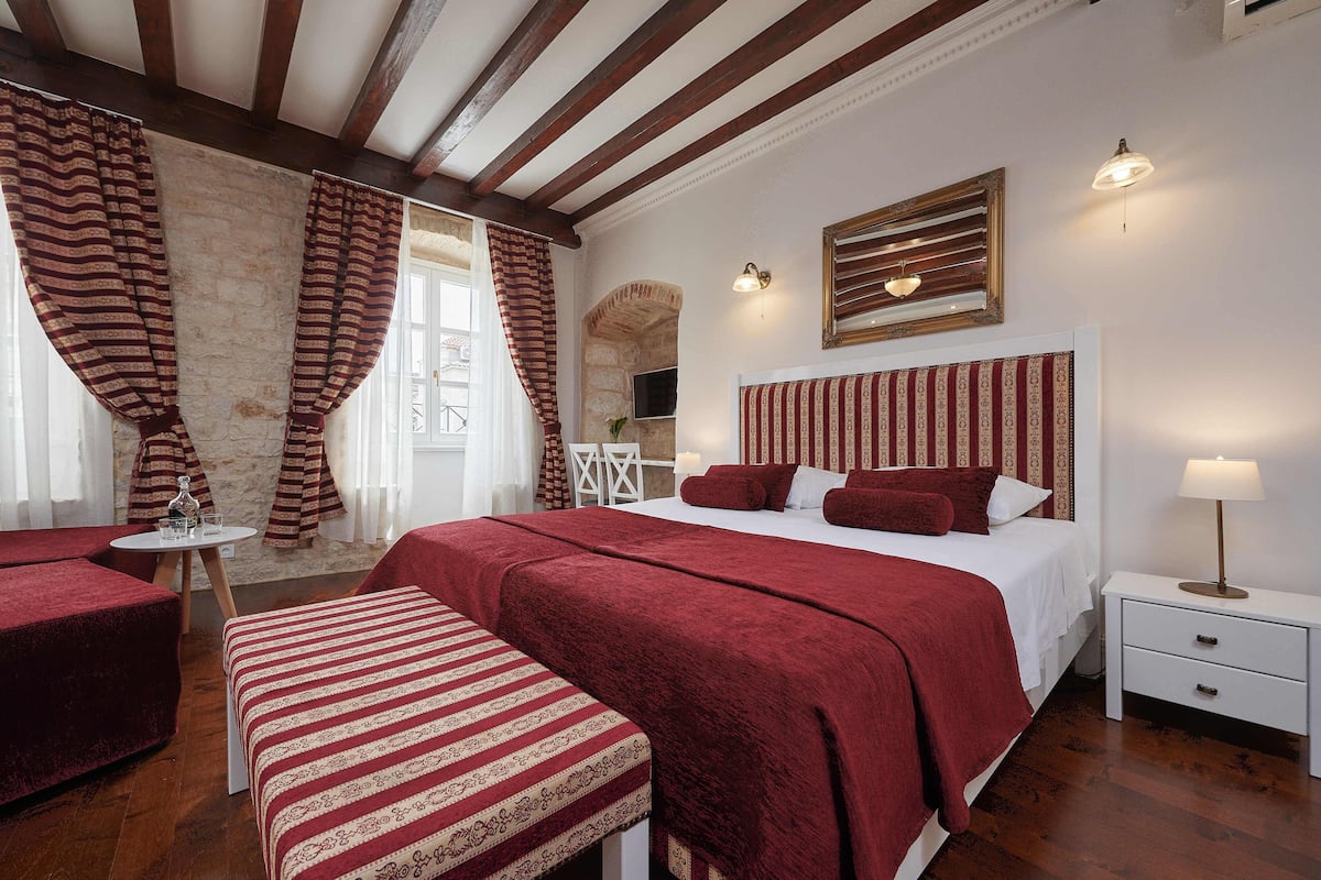 A bedroom with a large bed, a bedside table with a lamp, a small table with a glass and a bottle, and a bench.