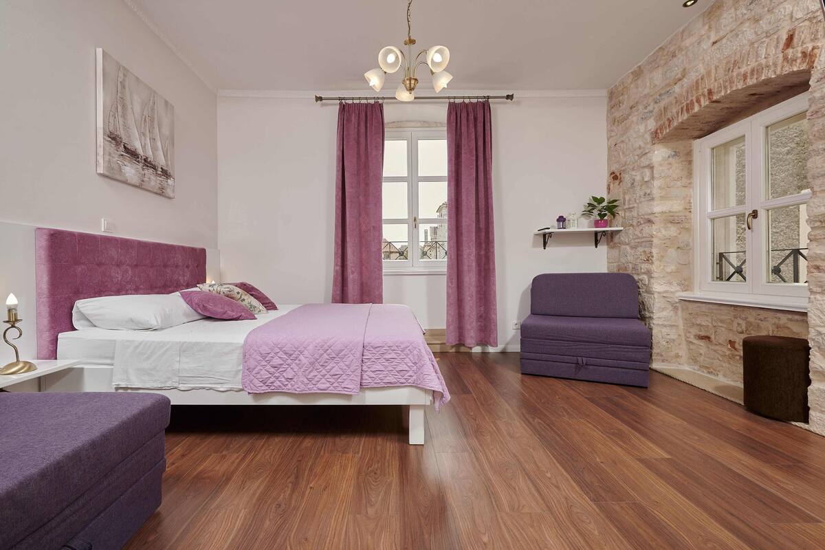 A bedroom with a bed, a chair, a window with curtains, a chandelier, and a stone wall.