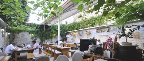 Un café en plein air avec des tables et des chaises en bois, entouré de verdure et de murs en pierre.