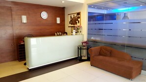 A reception area with a wooden reception desk, a clock, a framed artwork, a vase with flowers, and a brown sofa. - M Suites Hotel (Makati)