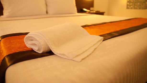 A neatly made bed with white linens and a rolled white towel on the side. - M Suites Hotel (Makati)