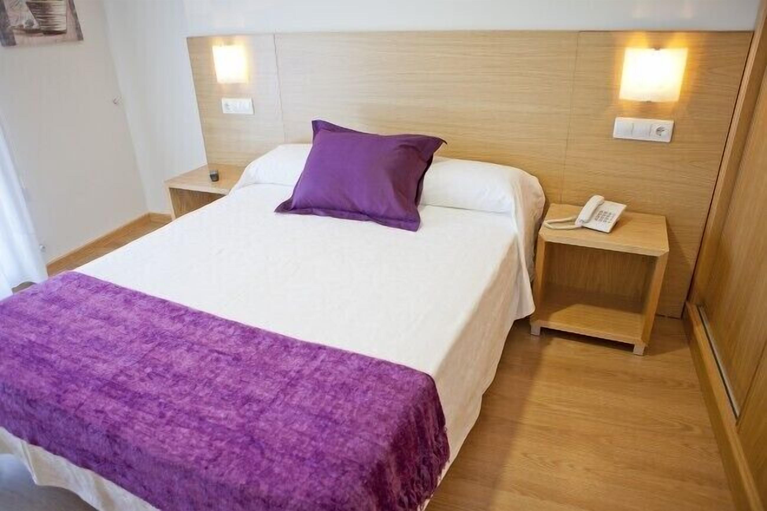 Habitación de hotel con cama, almohada morada, mesita de noche con teléfono y mesita auxiliar.