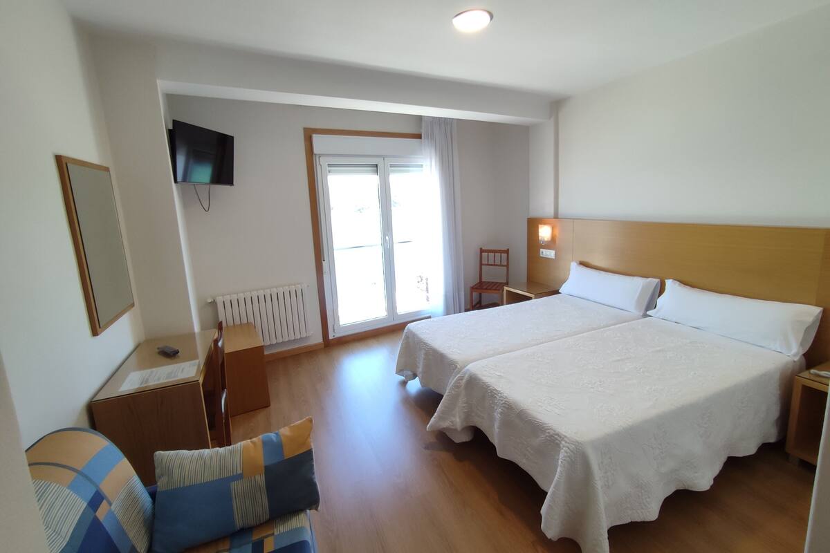 A hotel room with two beds, a television, a desk with a chair, a window with curtains, and a radiator.