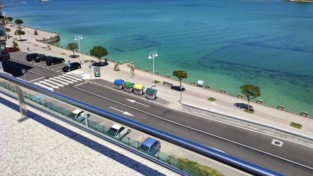 A coastal road with clear blue water, a pedestrian walkway, and parked cars. - Hotel Brisa del Mar (O Grove)