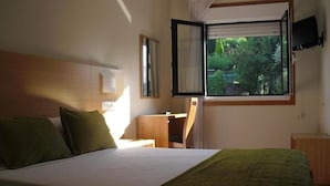 A hotel room with a bed, a desk, a chair, a TV, and a window with a view of greenery. - Hotel Brisa del Mar (O Grove)