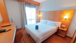 A hotel room with a large bed, wooden headboard, two bedside tables with lamps, a desk with a telephone, and a window with curtains.