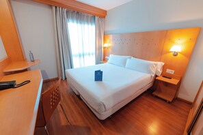 Habitación de hotel con una cama grande, cabecero de madera, dos mesitas de noche con lámparas, un escritorio con teléfono y una ventana con cortinas.