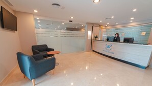 Zona de recepción de hotel con diseño moderno, que incluye un mostrador de recepción, un área de estar con un sofá y una mesita, y un espejo grande sobre el mostrador.