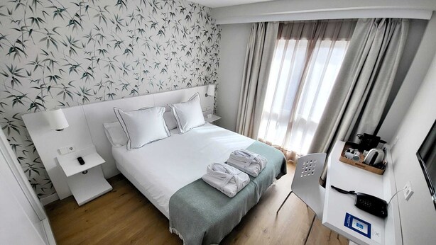 A hotel room with a bed, bedside table, lamp, and a desk with a phone and a kettle.