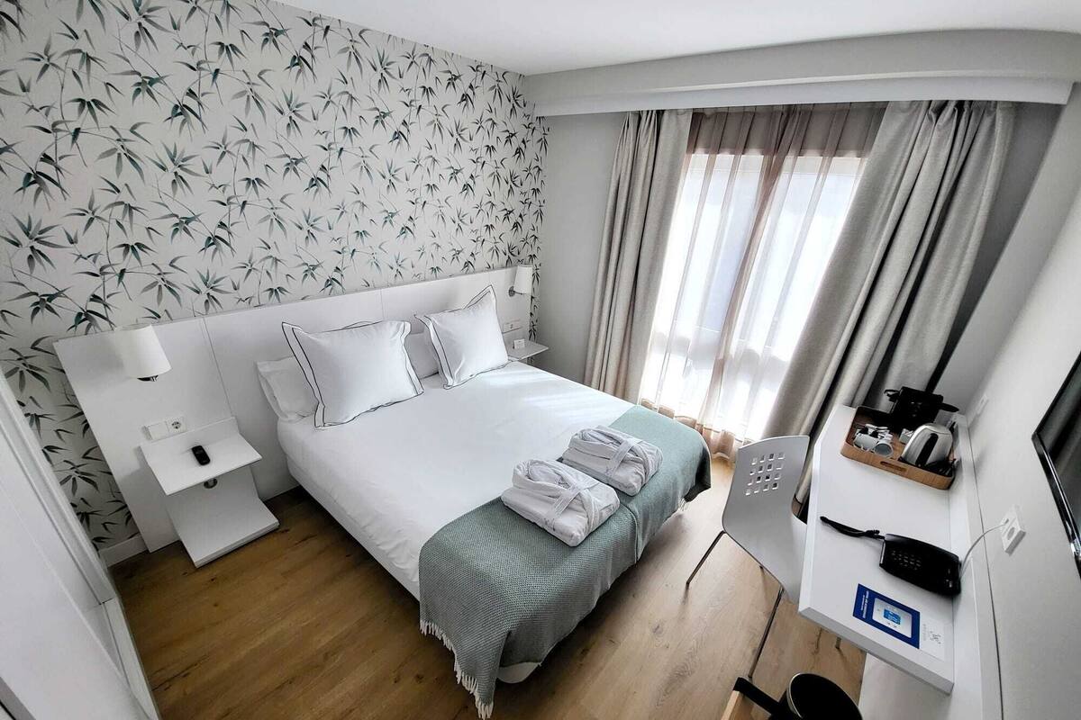 A hotel room with a bed, bedside table, lamp, and a desk with a phone and a kettle.