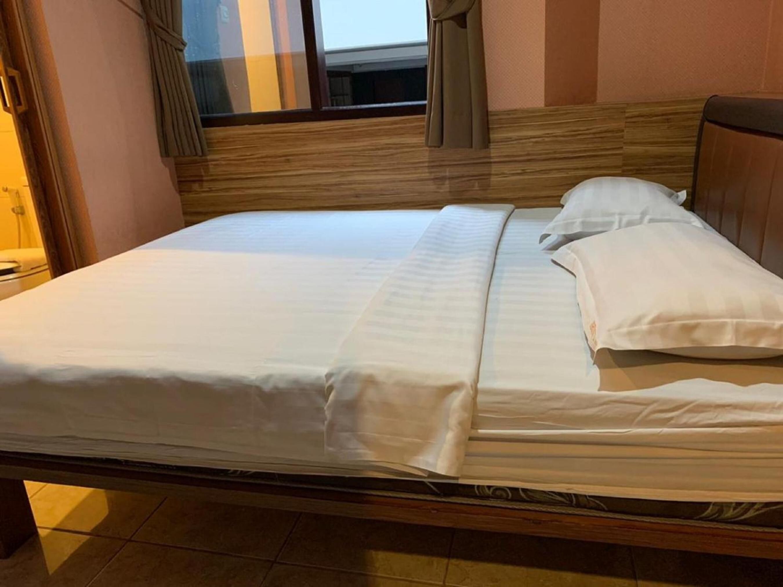 Een netjes opgemaakt bed met witte lakens in een kamer met een houten hoofdeinde en een raam met gordijnen.