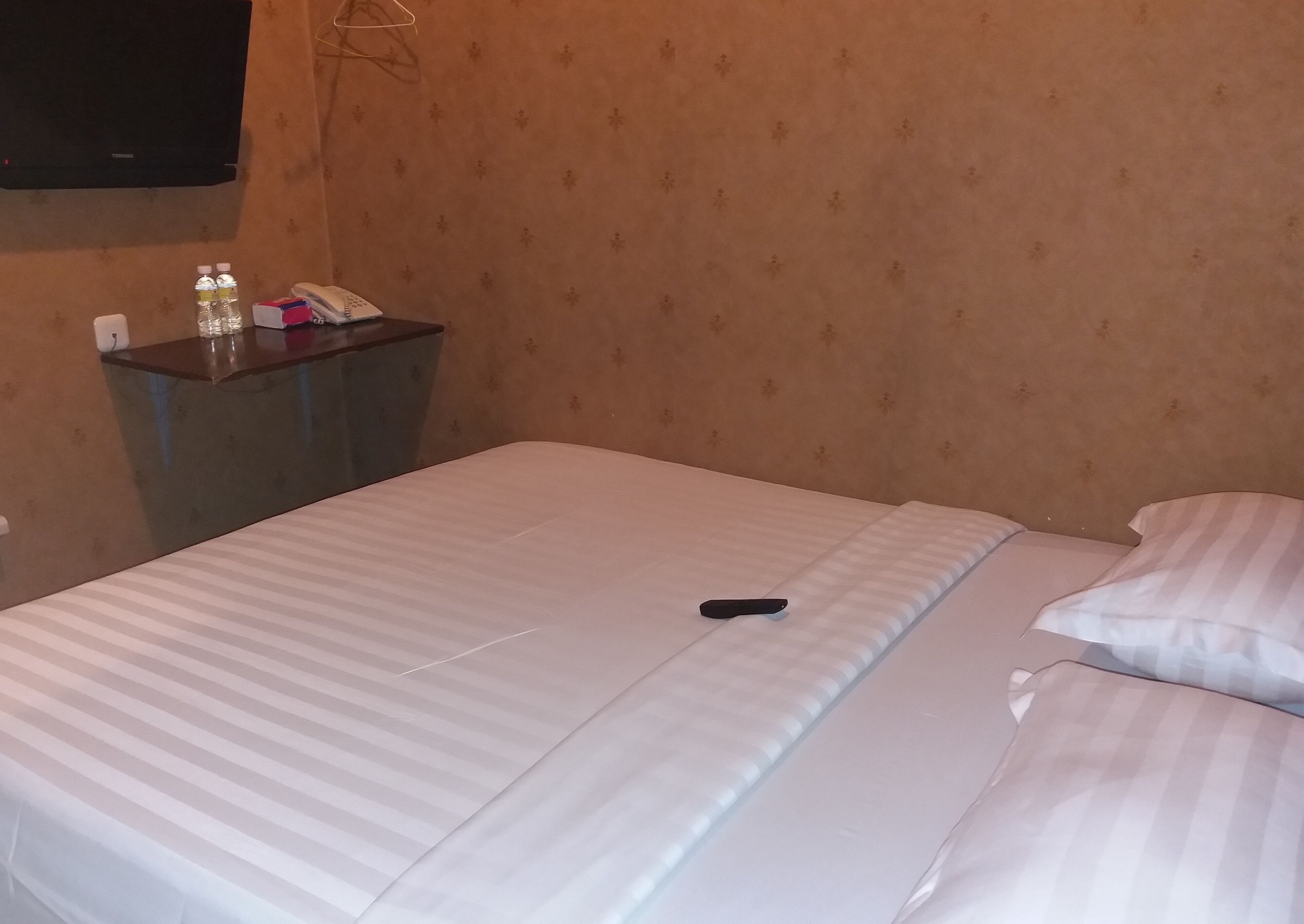 A hotel room with a bed, a bedside table with a phone and bottled water, and a wall-mounted TV.