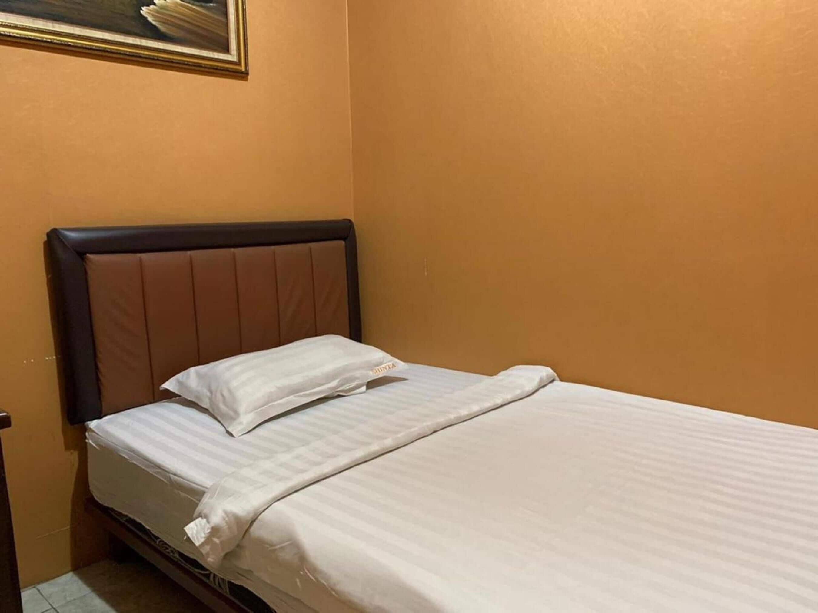 A single bed with a headboard, a white pillow, and a white bedspread in a room with orange walls.