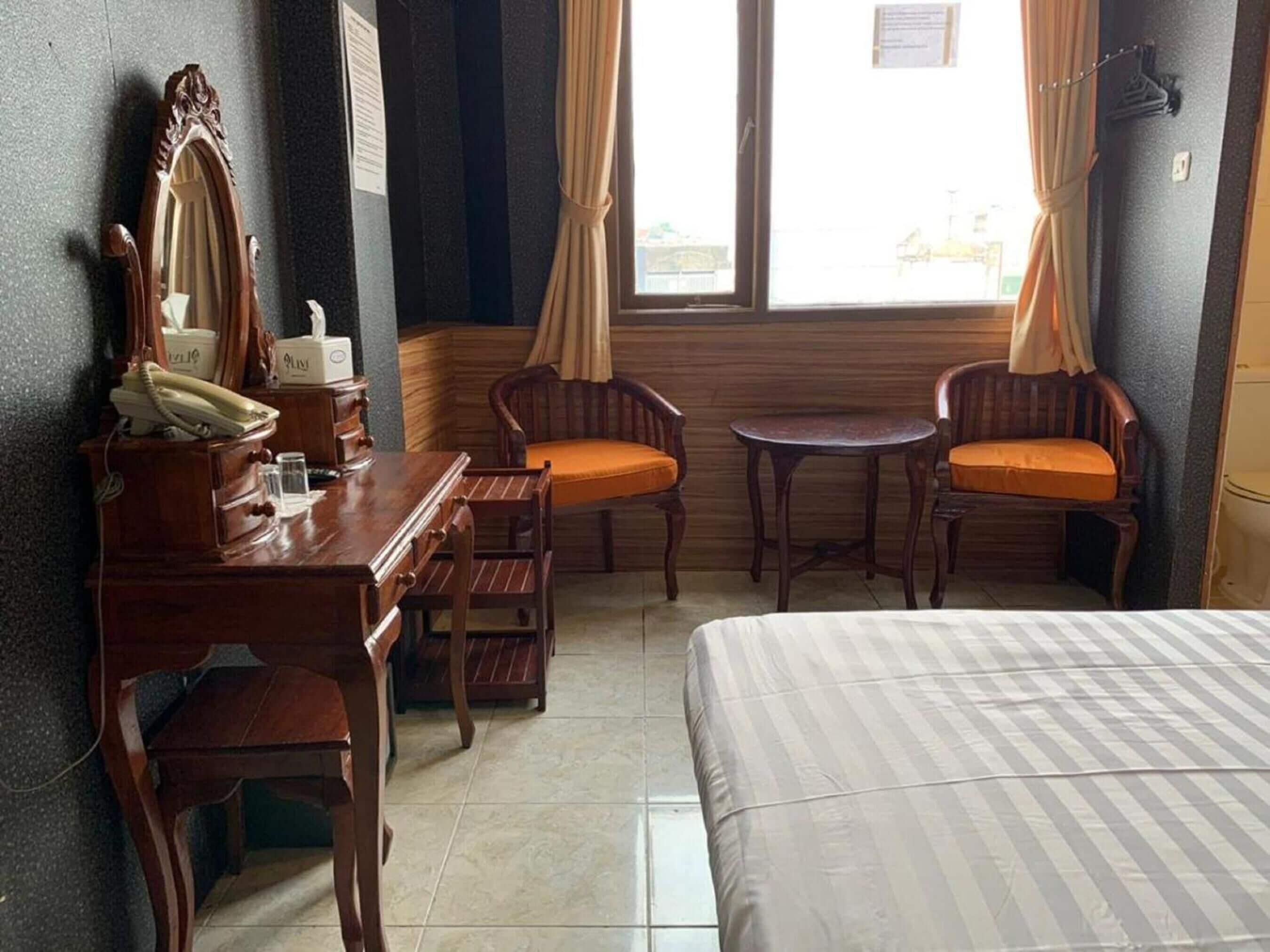 Habitación de hotel con cama, escritorio con teléfono, dos sillas, una mesita y una ventana con cortinas.