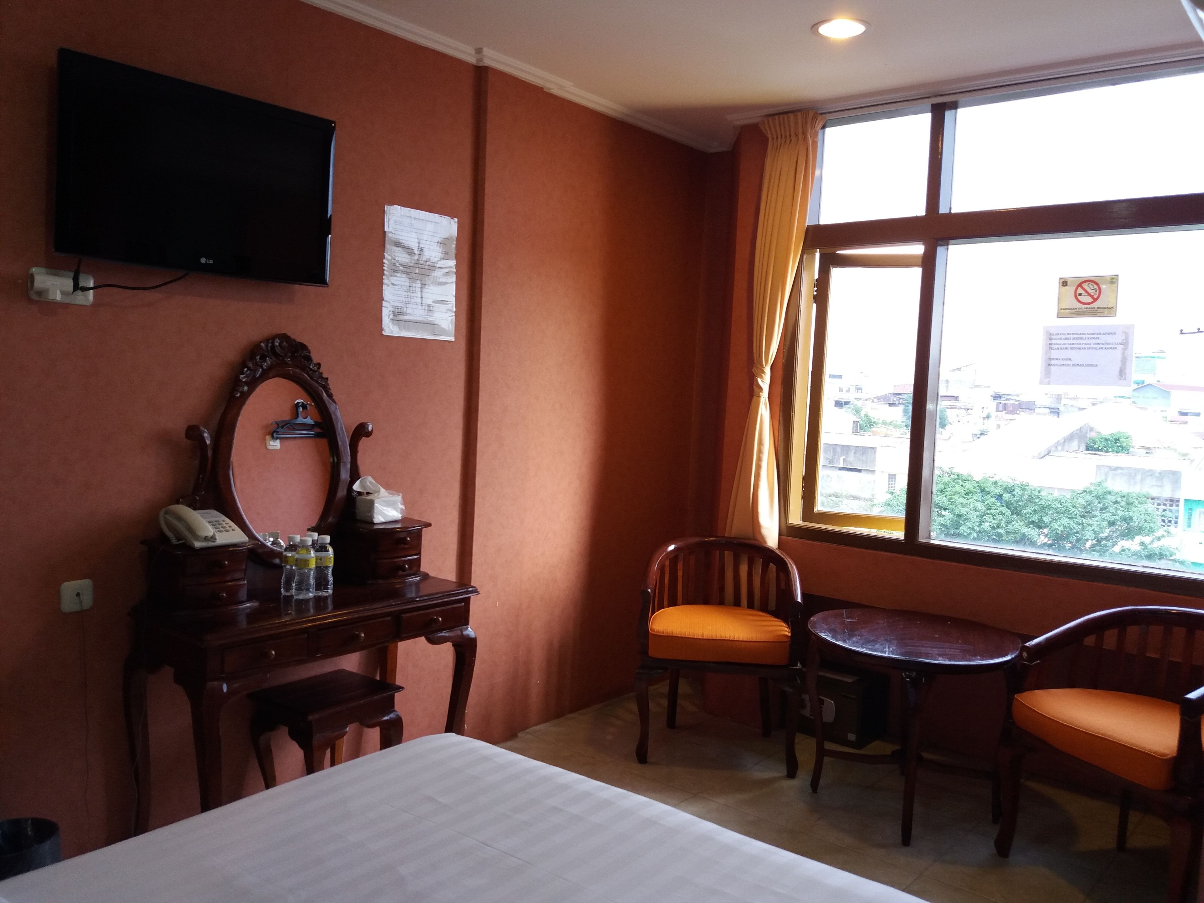 A hotel room with a bed, a desk with a telephone, a mirror, a television, and a window with a view of buildings.
