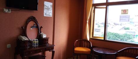 A hotel room with a bed, a desk with a telephone, a mirror, a television, and a window with a view of buildings.